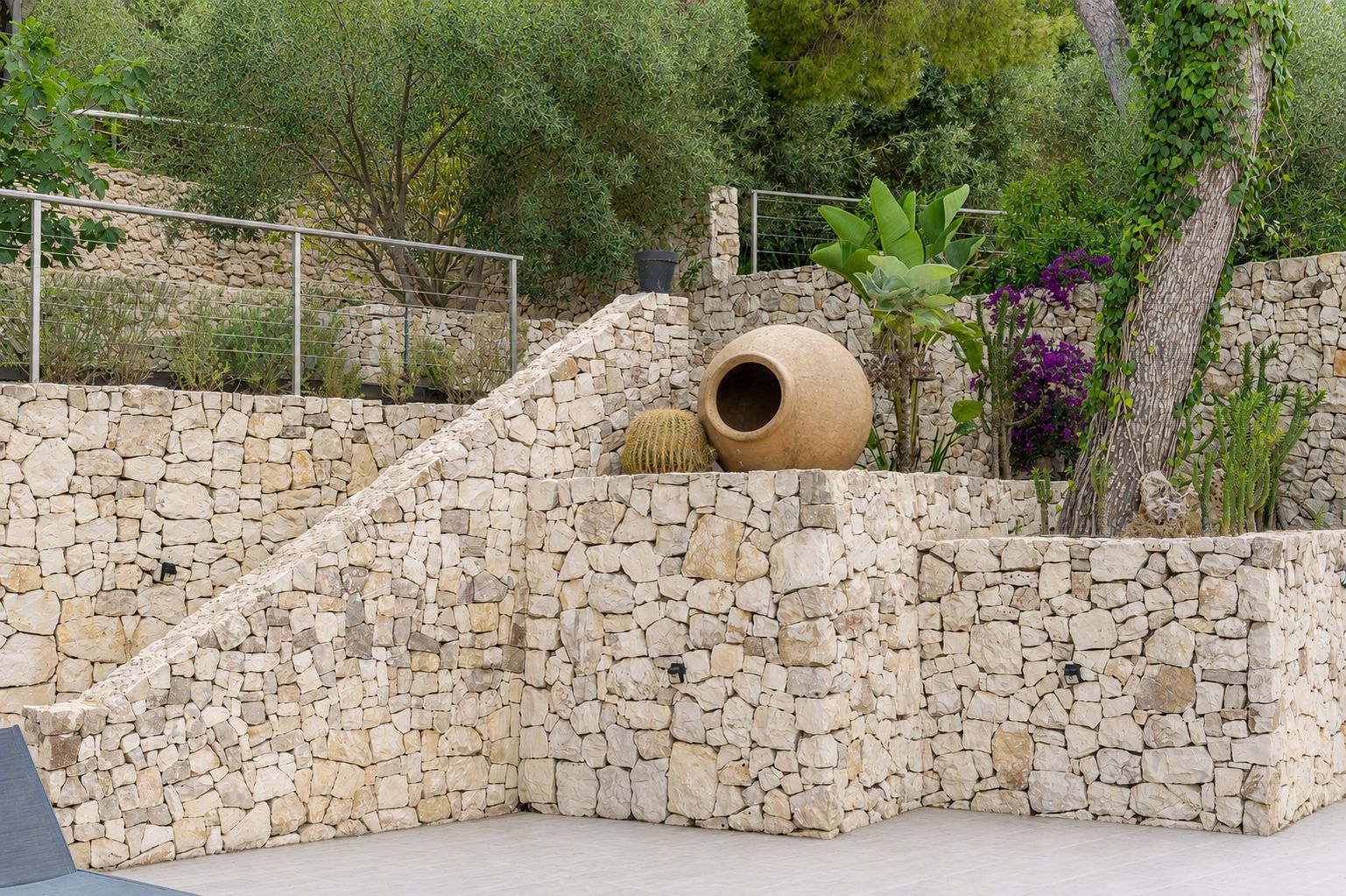 Zona de jardín aterrazado con muros de piedra seca, escaleras integradas y variada flora mediterránea. Una gran tinaja decorativa y cactus realzan la estética natural y rústica.