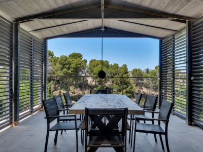 Amplia terraza cubierta con comedor exterior, mesa de madera rústica y sillas oscuras, enmarcada por celosías que ofrecen privacidad y vistas despejadas a la vegetación mediterránea.
