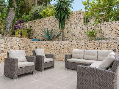 Amplia terraza exterior con mobiliario de ratán y cojines claros, enmarcada por muros de piedra seca escalonados y exuberante vegetación mediterránea.