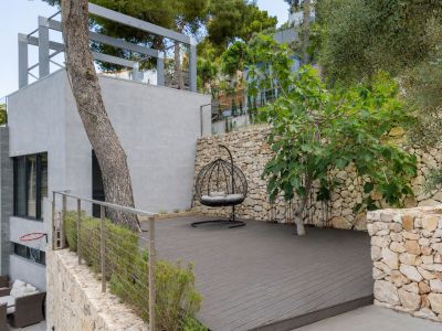 Amplia terraza exterior con tarima de composite oscuro, equipada con un sillón colgante y rodeada de muros de piedra natural. Árboles mediterráneos y arbustos aportan privacidad y se integran con la arquitectura moderna.