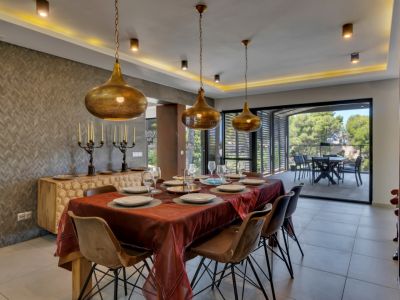 Amplio comedor con mesa de madera y sillas modernas, iluminado por tres lámparas colgantes doradas y luz indirecta. La pared texturizada y el aparador tallado añaden carácter, con acceso a una terraza exterior.