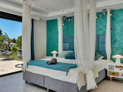 Amplio dormitorio con cama con dosel y cabecero tapizado azul, pared decorativa turquesa texturizada y acceso directo a terraza exterior con sillas colgantes y vegetación.