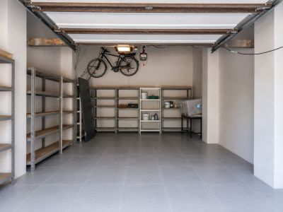 Amplio garaje con suelo de baldosas grises, extensas estanterías metálicas para almacenamiento organizado y soporte para bicicleta en pared. La estructura del techo expuesta ofrece un toque funcional.