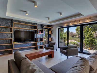Amplio salón con estanterías de madera a medida y TV integrada en pared texturizada. Grandes ventanales ofrecen vistas verdes, junto a cómodos sofás grises y suelo de baldosas claras.