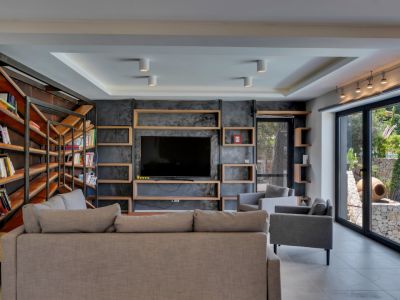 Amplio salón con pared de TV en acabado texturizado oscuro y estantes de madera, complementado por una estantería industrial y ventanales al jardín mediterráneo.