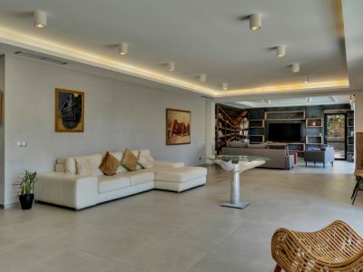 Amplio salón con sofá seccional blanco y mesa de diseño, conectado a una zona de biblioteca y TV con pared oscura y estanterías de madera.