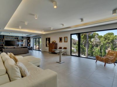Amplio salón de concepto abierto con grandes ventanales, ofreciendo vistas a la exuberante vegetación y luz natural. Destaca el sofá seccional blanco y la moderna iluminación empotrada.