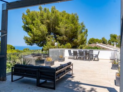 Amplio solárium en la azotea con moderno sofá exterior y conjunto de comedor, barandillas de cristal y vistas panorámicas al mar entre pinos mediterráneos.