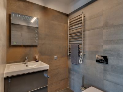 Aseo de cortesía con lavabo suspendido sobre mueble gris, espejo iluminado y toallero calefactado. Destacan los azulejos texturizados en tonos grises y óxido.