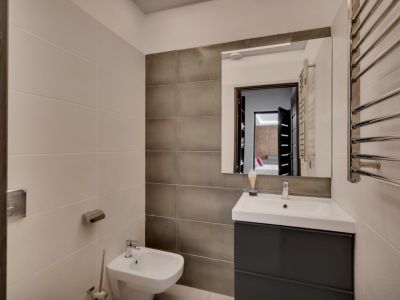 Baño contemporáneo con mueble de lavabo gris oscuro, lavamanos blanco, bidé y toallero calefactado cromado. Las paredes combinan azulejos claros texturizados y oscuros lisos.