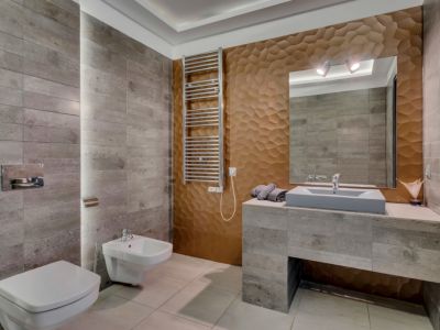 Baño moderno con azulejos grises texturizados, una pared decorativa 3D geométrica en tono bronce y un lavabo flotante. Incluye inodoro y bidé suspendidos, además de toallero calefactado.