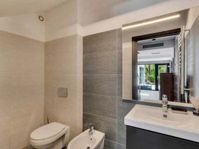 Baño moderno con azulejos texturizados en tonos neutros, inodoro, bidé y lavabo con mueble oscuro. El espejo iluminado refleja un exterior ajardinado.