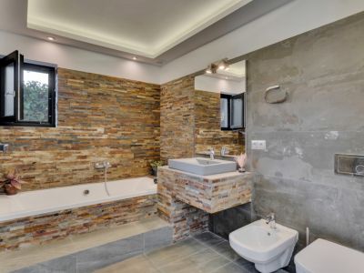 Baño moderno con bañera y lavabo flotante revestidos en piedra apilada multicolor. Grandes azulejos grises cubren paredes y suelo, con iluminación empotrada.