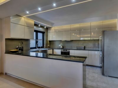 Cocina moderna con armarios de alto brillo color crema, encimeras de granito negro y electrodomésticos integrados. Iluminación LED lineal y empotrada en el techo.