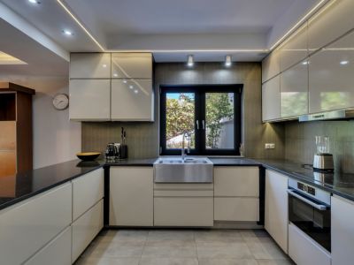 Cocina moderna con armarios lacados en blanco roto, encimeras negras y fregadero de acero inoxidable bajo una ventana que da a la vegetación. Iluminación LED integrada y suelo de baldosas grises.