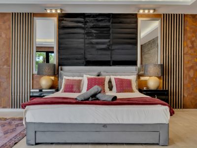 Dormitorio principal con cama tapizada gris, cabecero negro texturizado, espejos y paneles de pared únicos en madera, metal envejecido y piedra. Iluminación a medida crea un ambiente sofisticado.
