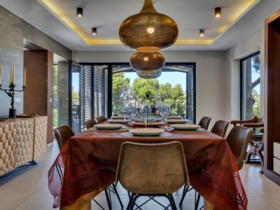 Elegante comedor con lámparas colgantes de bronce ornamentadas, un aparador de madera texturizada y amplios ventanales con vistas serenas a la exuberante vegetación.