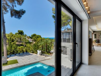 Grandes puertas correderas de cristal negro conectan el interior moderno con la piscina privada, terraza de gres y exuberante paisaje mediterráneo con vistas al mar lejano.