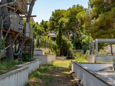 Jardín con una casa en el árbol de madera rústica entre pinos maduros y una hamaca. El espacio incluye muros de contención, vegetación densa y una pérgola moderna.