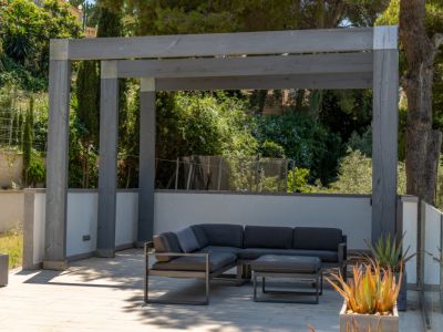 Moderna zona de estar exterior bajo una pérgola de madera gris, con sofá en L y otomana sobre pavimento claro, rodeada de vegetación mediterránea y plantas suculentas en macetas.