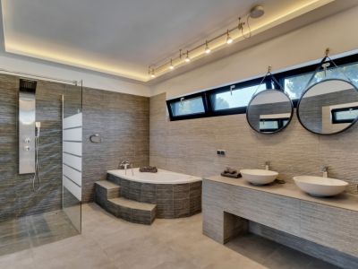 Moderno baño con ducha de hidromasaje, bañera esquinera con escalones y doble lavabo. Azulejos texturizados, espejos redondos y luz natural de ventanas horizontales.