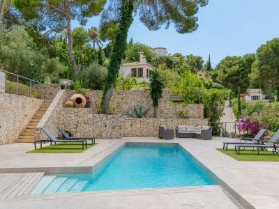 Piscina privada rectangular con escalones integrados, rodeada de pavimento claro y zonas de césped artificial con tumbonas. Muros de piedra natural y vegetación mediterránea en terrazas ajardinadas.