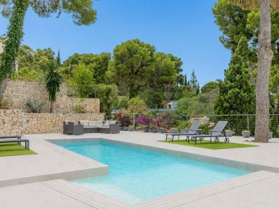 Piscina privada rectangular rodeada de una amplia terraza de gres claro, con zona de relax exterior y muro de piedra natural en un entorno ajardinado.