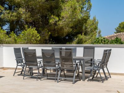 Terraza exterior con mesa de comedor para ocho personas y sillas de diseño moderno, sobre suelo de baldosas claras, enmarcada por una pared blanca y vistas a la vegetación y tejados tradicionales.