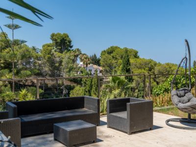 Terraza exterior con mobiliario de ratán oscuro y silla colgante, ofreciendo vistas panorámicas a la exuberante vegetación y una casa tradicional en la distancia.