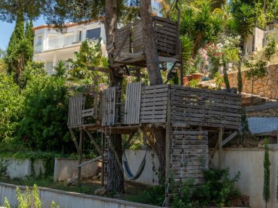 Una rústica casa en el árbol de varios niveles, construida con palets de madera, con escaleras y una hamaca, rodeada de vegetación y una villa moderna al fondo.