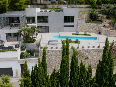 Vista aérea de la villa moderna con piscina rectangular y terrazas escalonadas. Destacan la fachada gris, los jardines en bancales de piedra y las amplias zonas de ocio exterior.
