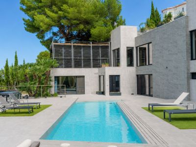 Vista exterior de la villa con piscina privada rectangular, terrazas de gres gris y zonas de césped artificial. Destaca su arquitectura moderna y una terraza superior con lamas.