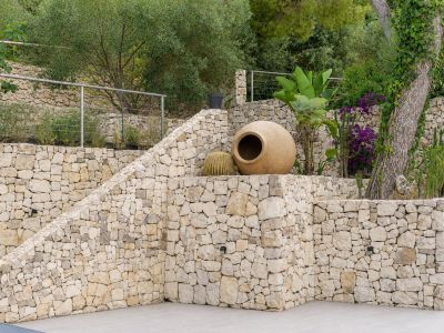 Zona de jardín aterrazado con muros de piedra seca, escaleras integradas y variada flora mediterránea. Una gran tinaja decorativa y cactus realzan la estética natural y rústica.