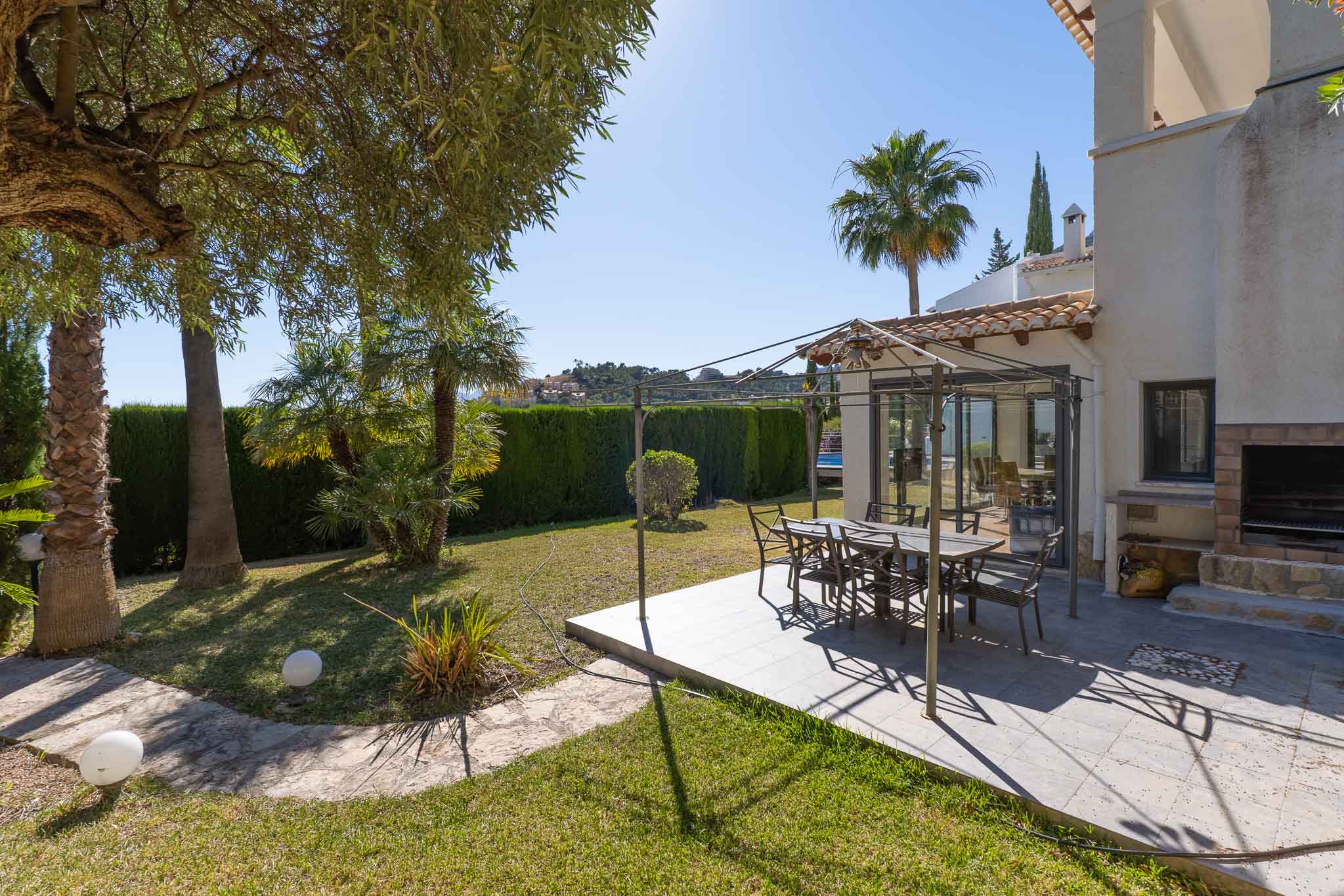 Acogedor comedor exterior con pérgola metálica sobre patio embaldosado, ideal para comidas al aire libre. Se complementa con una barbacoa de obra y un frondoso jardín con palmeras y seto de privacidad. Acogedor comedor exterior con pérgola metálica sobre patio embaldosado, ideal para comidas al aire libre. Se complementa con una barbacoa de obra y un frondoso jardín con palmeras y seto de privacidad.