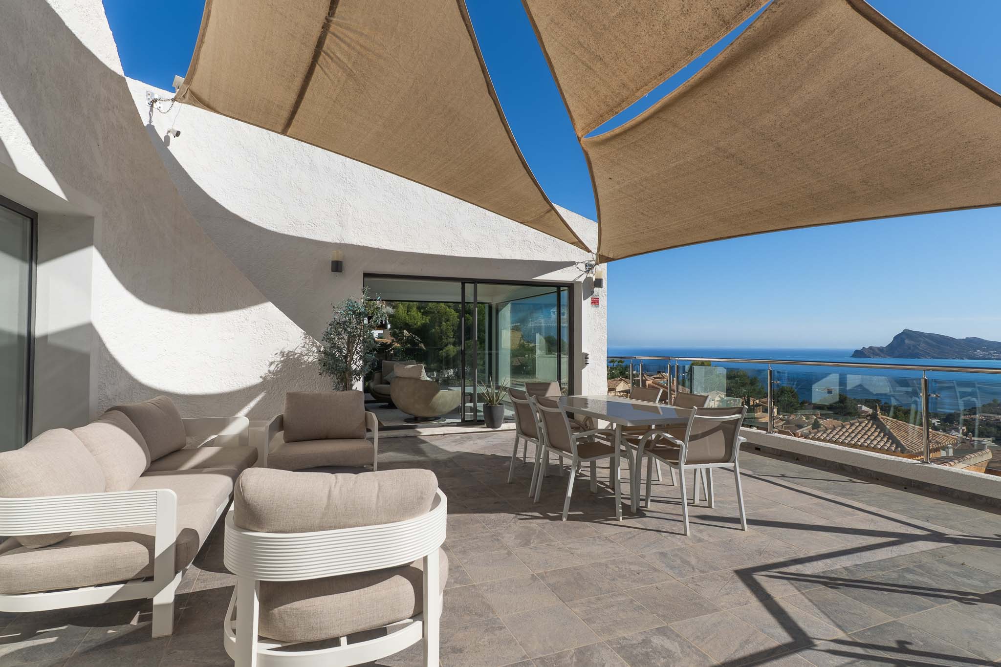 Amplia terraza exterior con zona de estar y comedor bajo vela de sombra, ofreciendo vistas panorámicas al mar y la costa a través de barandilla de cristal. Amplia terraza exterior con zona de estar y comedor bajo vela de sombra, ofreciendo vistas panorámicas al mar y la costa a través de barandilla de cristal.