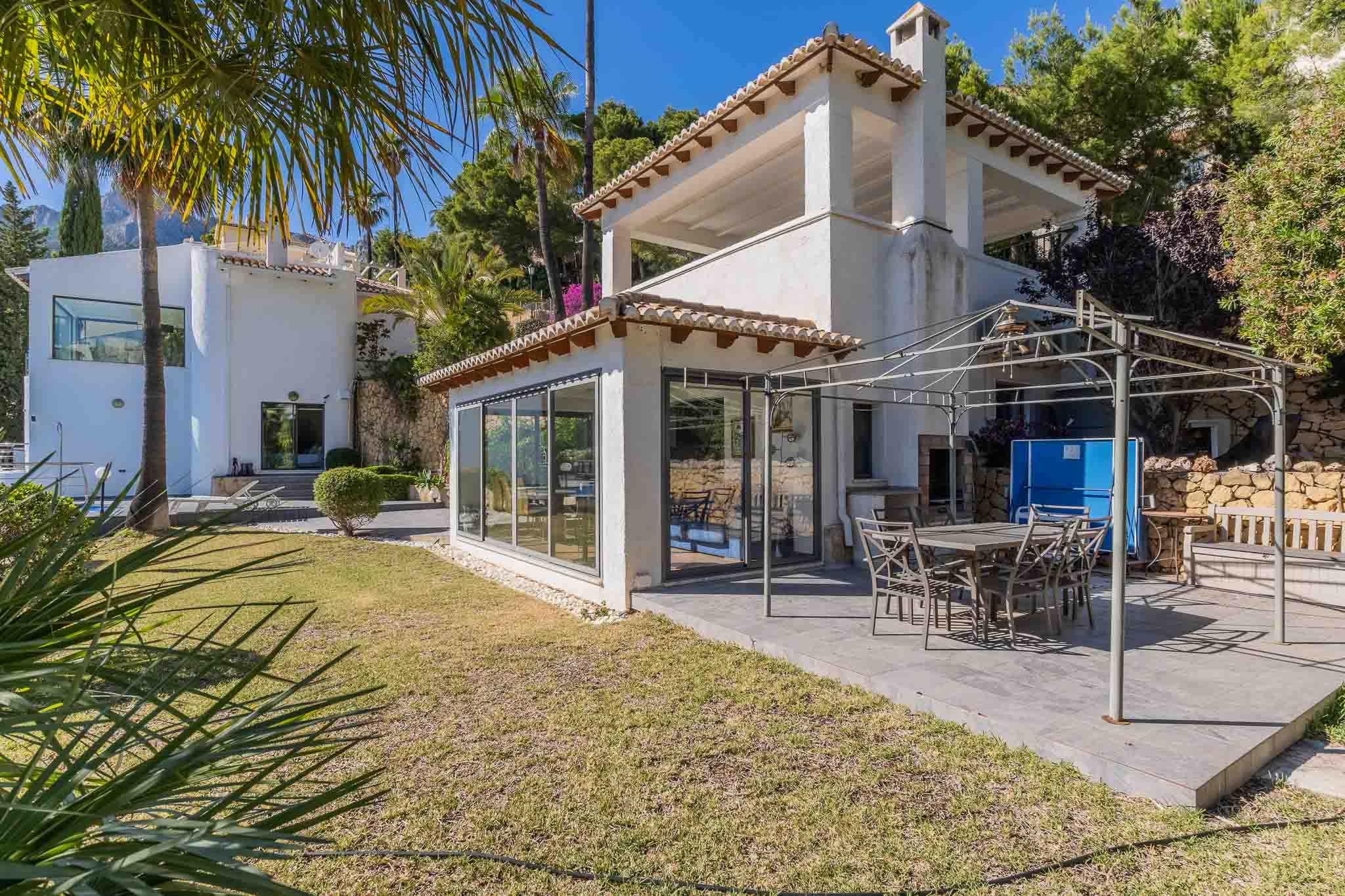 Amplia terraza pavimentada con comedor exterior bajo pérgola metálica, adyacente a una sala acristalada y rodeada de un cuidado jardín mediterráneo. Amplia terraza pavimentada con comedor exterior bajo pérgola metálica, adyacente a una sala acristalada y rodeada de un cuidado jardín mediterráneo.