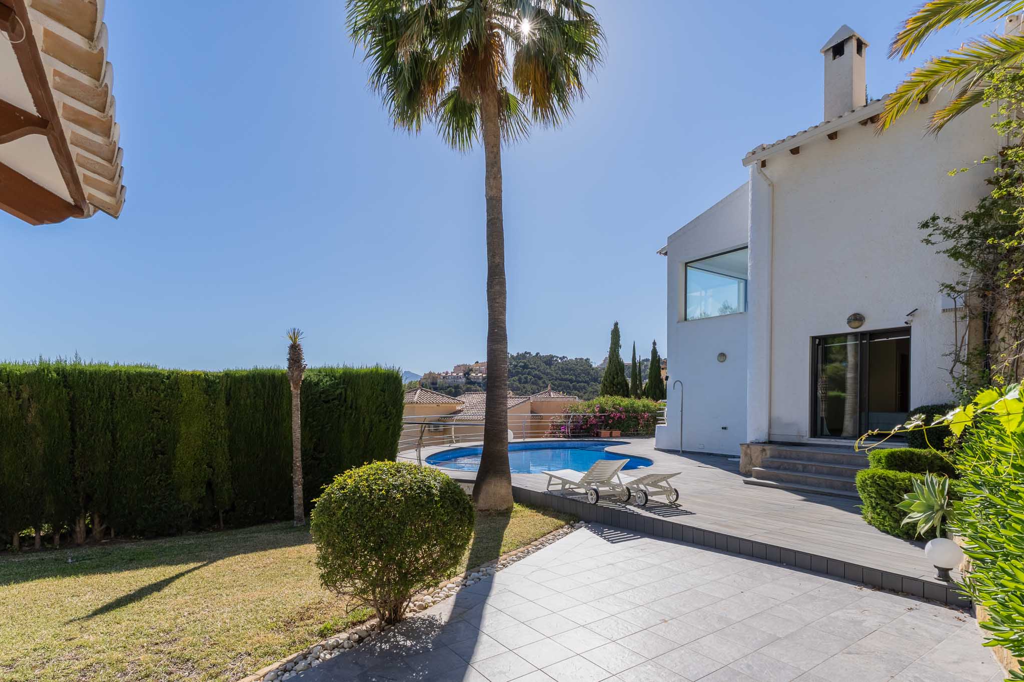 Amplio jardín con piscina circular y solárium de tarima, flanqueado por una villa blanca y un gran seto que ofrece privacidad. Vistas a las colinas. Amplio jardín con piscina circular y solárium de tarima, flanqueado por una villa blanca y un gran seto que ofrece privacidad. Vistas a las colinas.