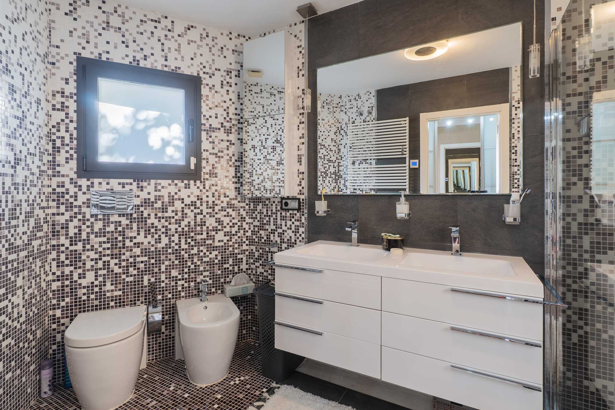 Baño con paredes y suelo de mosaico en tonos tierra y gris. Destacan el doble lavabo blanco con mueble suspendido, un gran espejo iluminado y sanitarios murales, complementados por un radiador toallero. Baño con paredes y suelo de mosaico en tonos tierra y gris. Destacan el doble lavabo blanco con mueble suspendido, un gran espejo iluminado y sanitarios murales, complementados por un radiador toallero.