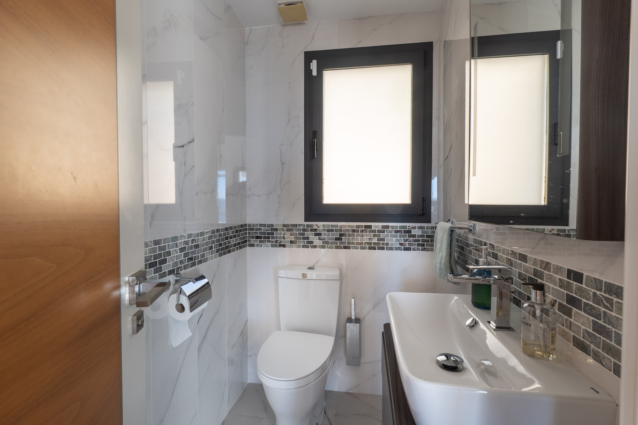 Baño moderno con azulejos efecto mármol blanco, una banda decorativa de mosaico gris y ventana con marco oscuro. Incluye lavabo blanco y sanitarios a juego. Baño moderno con azulejos efecto mármol blanco, una banda decorativa de mosaico gris y ventana con marco oscuro. Incluye lavabo blanco y sanitarios a juego.