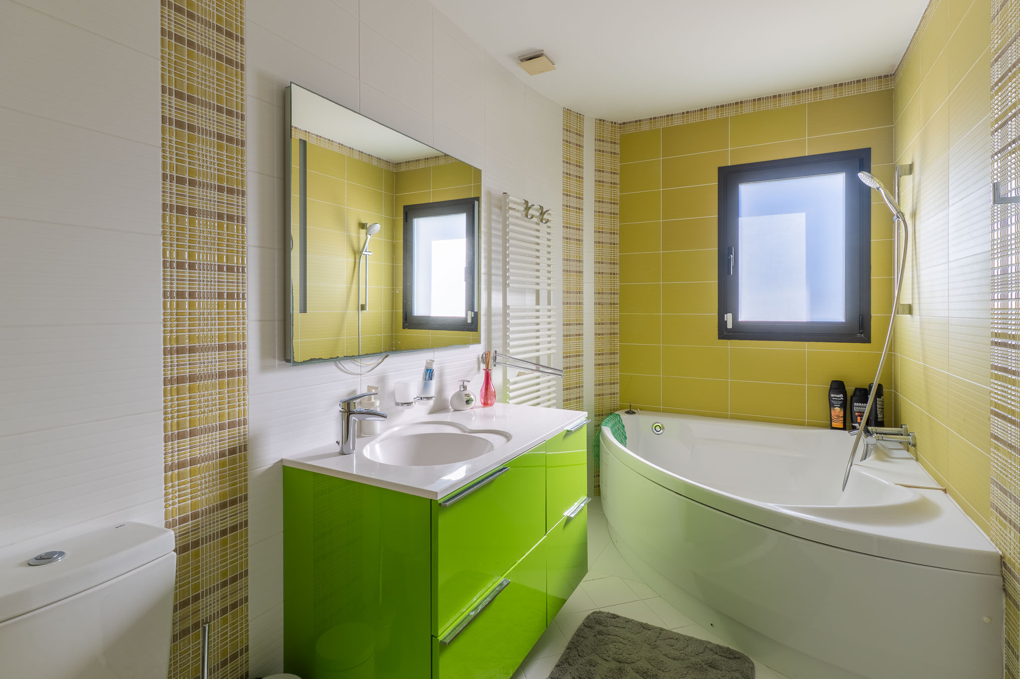 Baño moderno con bañera de esquina y lavabo verde brillante. Azulejos blancos texturizados y verdes lima, con un detalle de mosaico a cuadros. Ventana con marco oscuro y toallero calefactable. Baño moderno con bañera de esquina y lavabo verde brillante. Azulejos blancos texturizados y verdes lima, con un detalle de mosaico a cuadros. Ventana con marco oscuro y toallero calefactable.