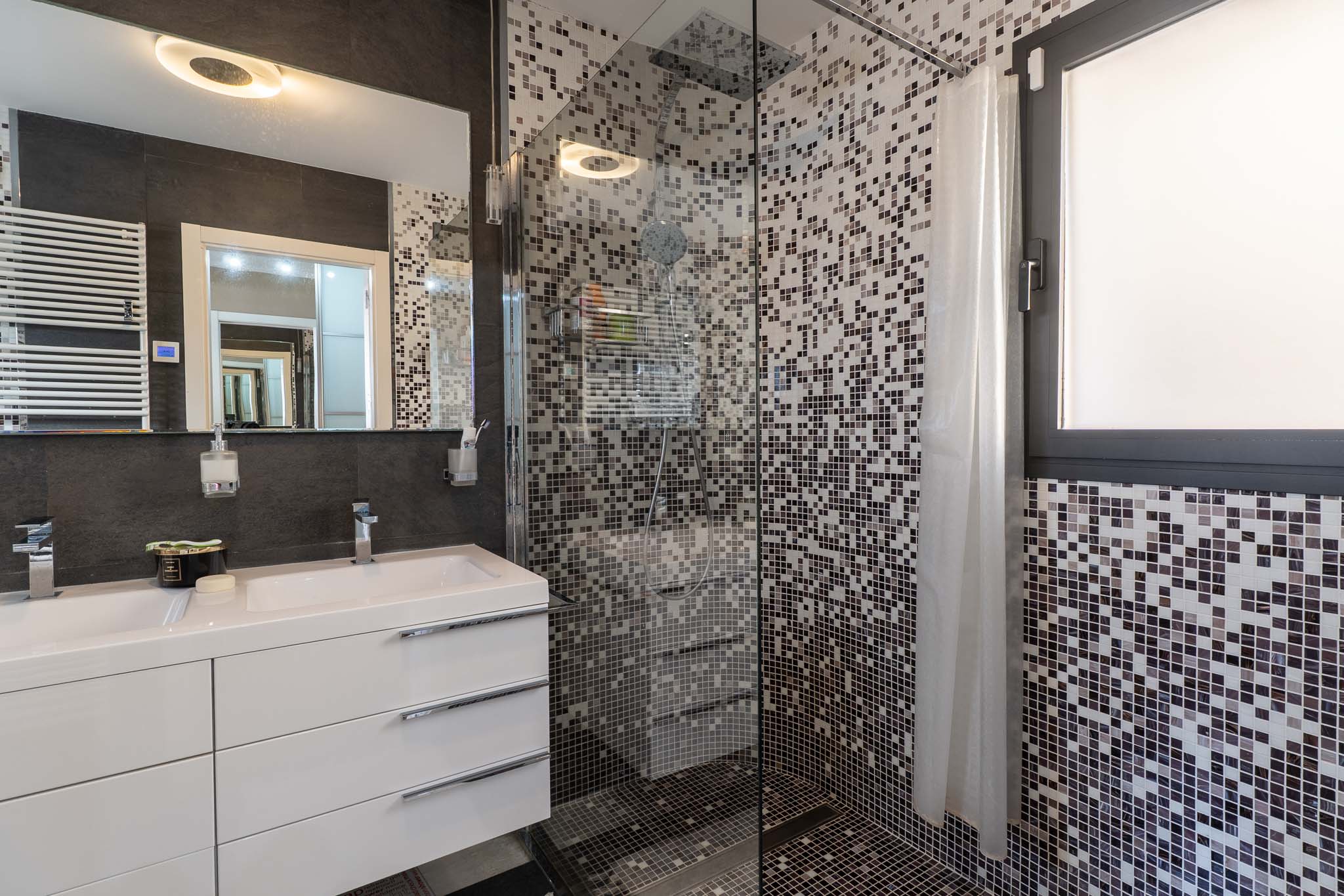 Baño moderno con doble lavabo, mueble blanco y espejo iluminado. La ducha de cristal presenta mosaico en tonos grises y una ventana integrada. Baño moderno con doble lavabo, mueble blanco y espejo iluminado. La ducha de cristal presenta mosaico en tonos grises y una ventana integrada.