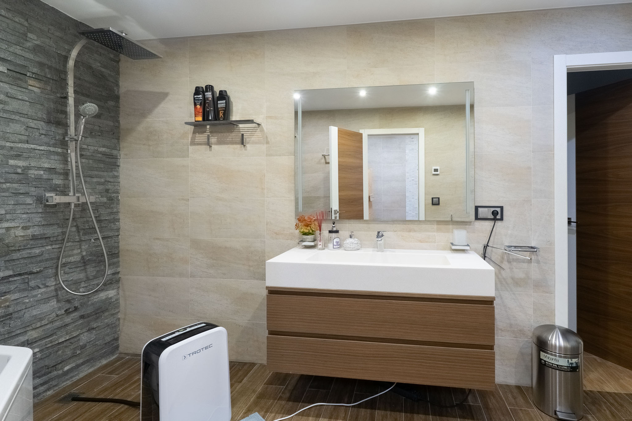 Baño moderno con ducha de lluvia y pared de pizarra, tocador flotante de madera y lavabo integrado. Suelo de baldosas imitación madera. Baño moderno con ducha de lluvia y pared de pizarra, tocador flotante de madera y lavabo integrado. Suelo de baldosas imitación madera.