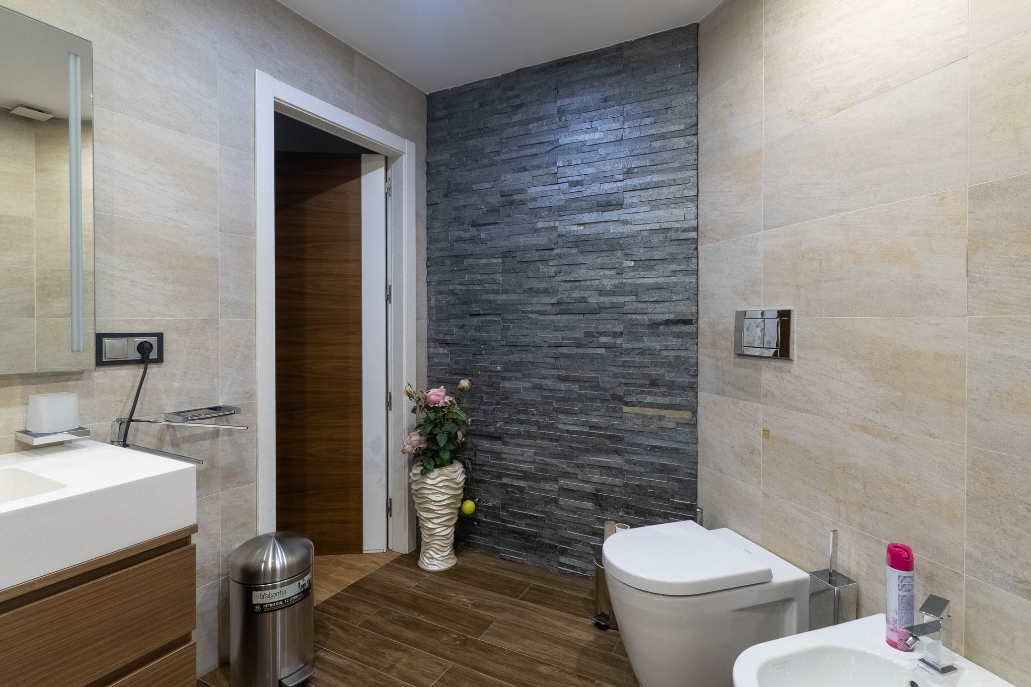 Baño moderno con pared decorativa de piedra apilada oscura, azulejos beige claro y suelo de imitación madera. Incluye inodoro, bidé y tocador con lavabo. Baño moderno con pared decorativa de piedra apilada oscura, azulejos beige claro y suelo de imitación madera. Incluye inodoro, bidé y tocador con lavabo.
