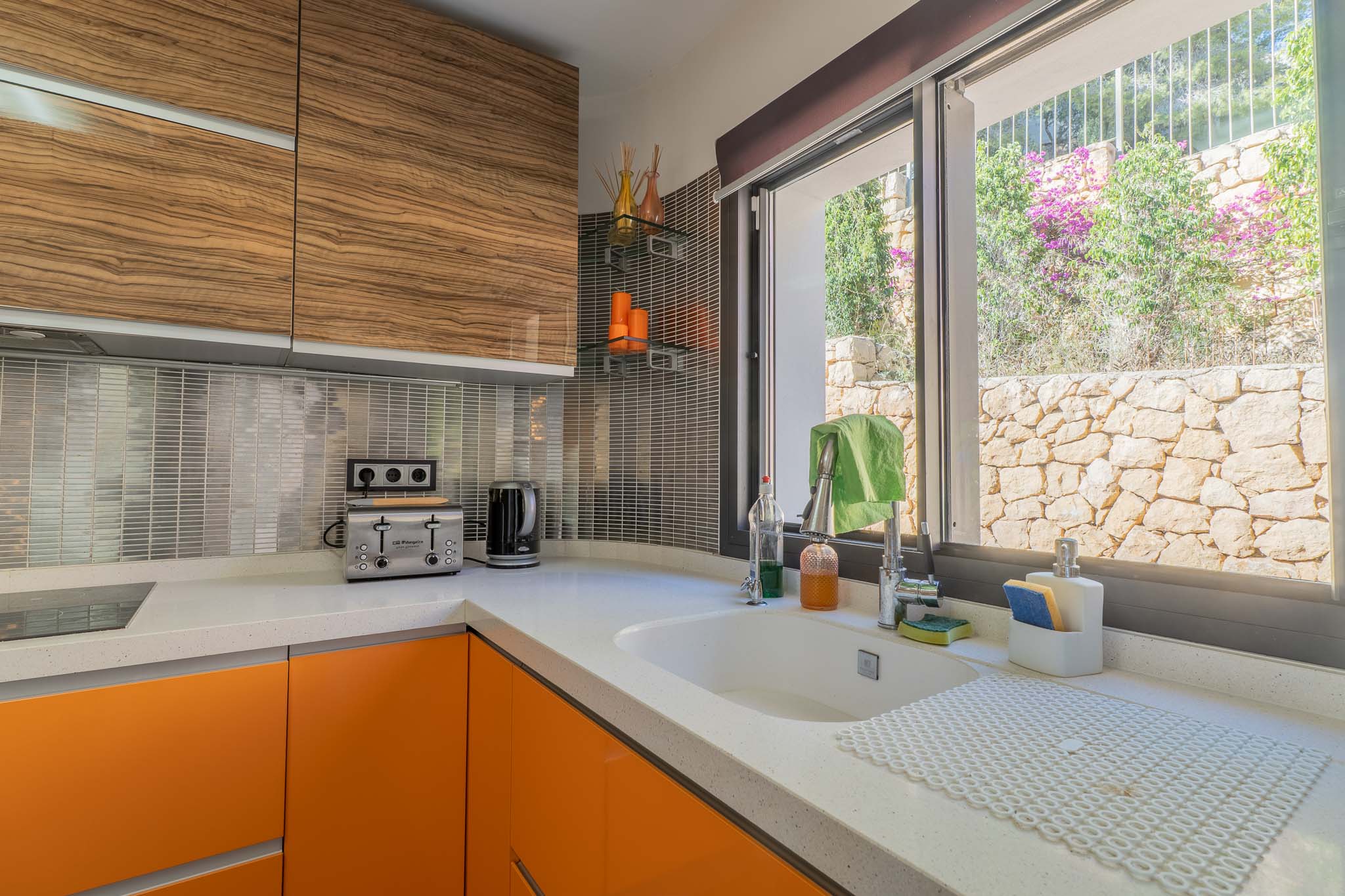 Cocina moderna con encimera de cuarzo blanco, armarios bajos naranjas y superiores de madera veteada. Ventana con vistas a un muro de piedra natural y vegetación. Cocina moderna con encimera de cuarzo blanco, armarios bajos naranjas y superiores de madera veteada. Ventana con vistas a un muro de piedra natural y vegetación.