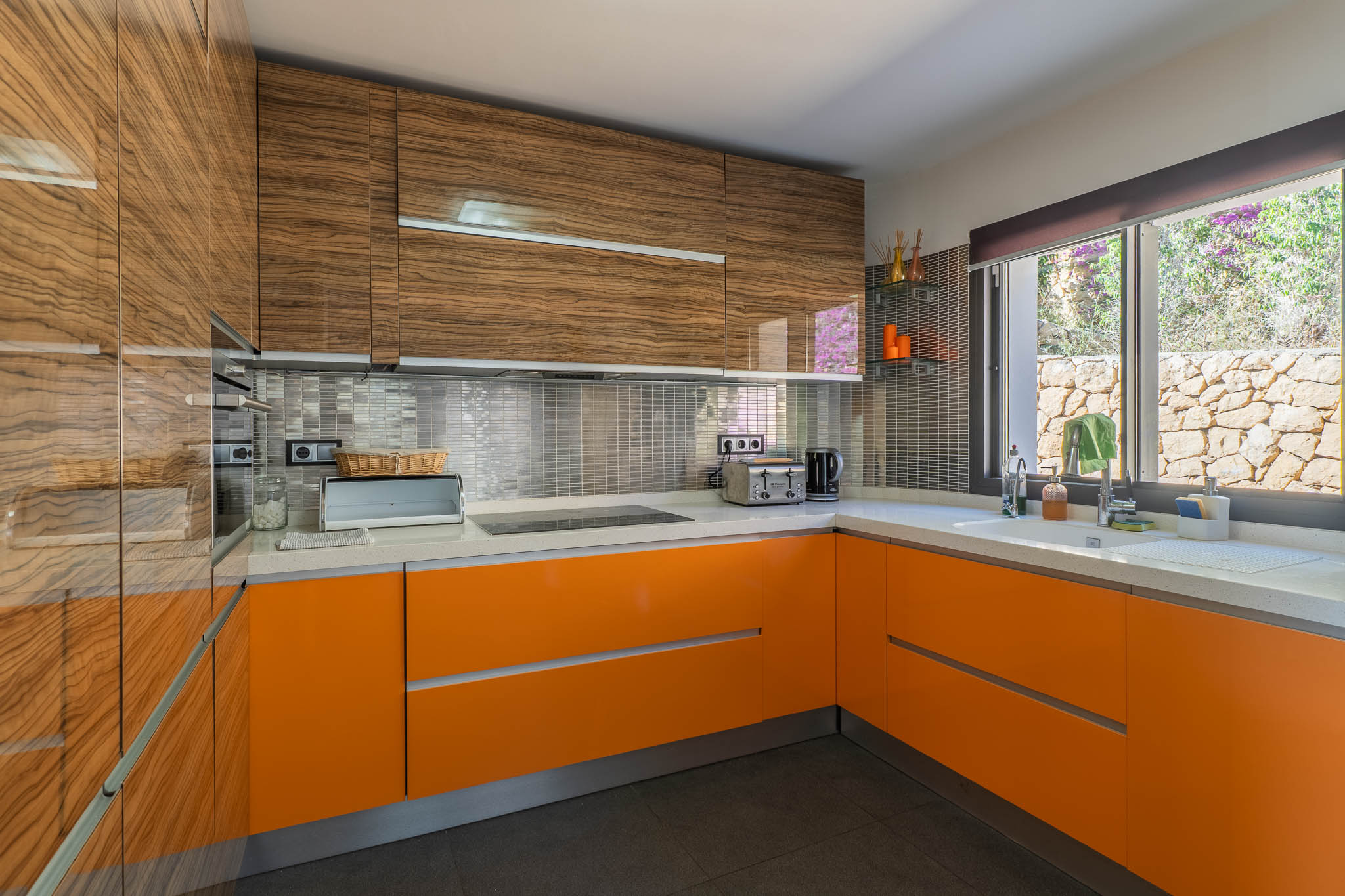 Cocina moderna con mobiliario superior de madera veteada y bajos lacados en naranja brillante. Destacan la encimera blanca, el salpicadero de mosaico metálico y la ventana con vistas a un muro de piedra. Cocina moderna con mobiliario superior de madera veteada y bajos lacados en naranja brillante. Destacan la encimera blanca, el salpicadero de mosaico metálico y la ventana con vistas a un muro de piedra.