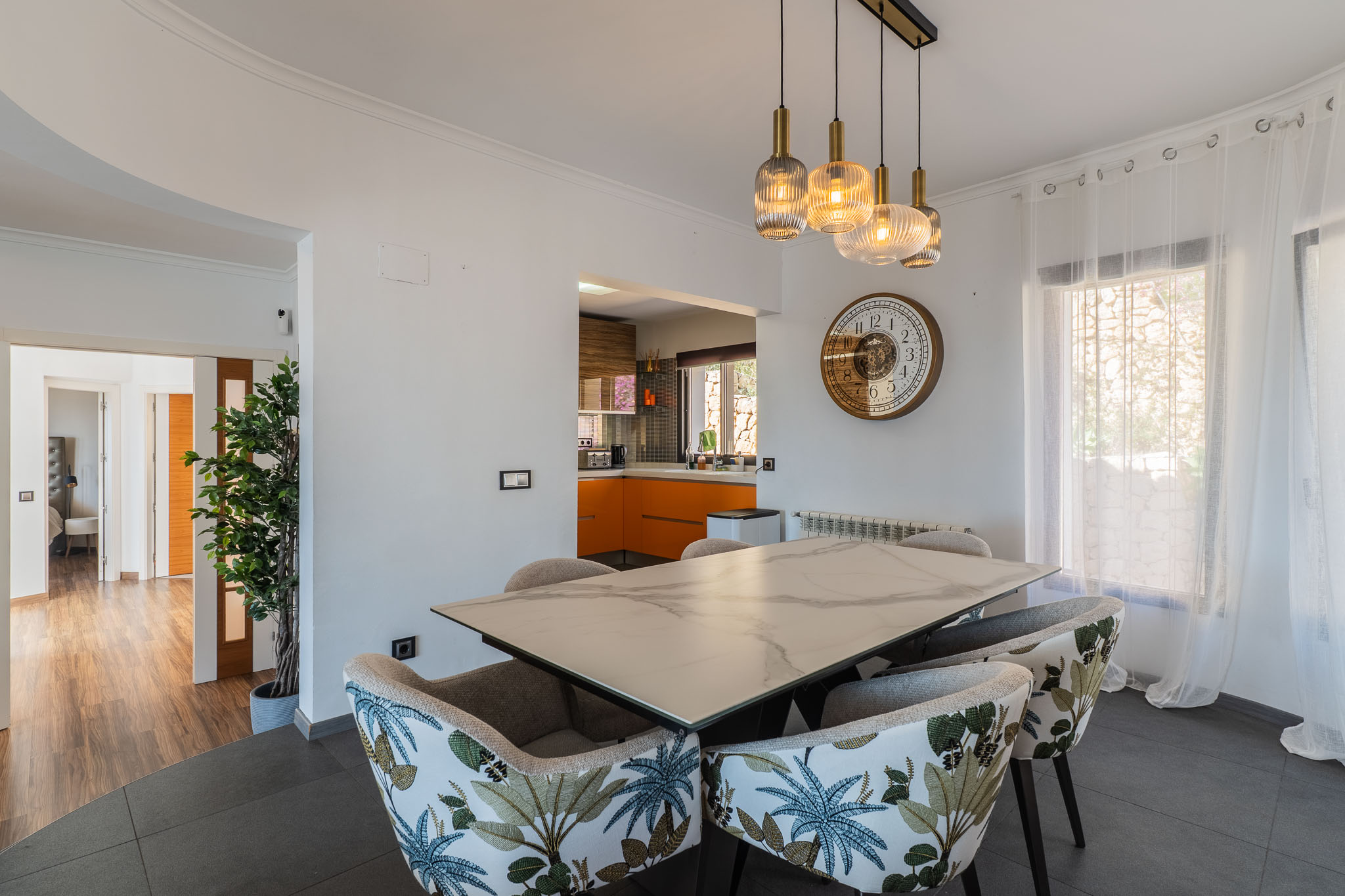 Comedor con mesa de efecto mármol y sillas tapizadas con patrón tropical. La iluminación colgante moderna ilumina el espacio, abierto a una cocina con gabinetes naranjas. Comedor con mesa de efecto mármol y sillas tapizadas con patrón tropical. La iluminación colgante moderna ilumina el espacio, abierto a una cocina con gabinetes naranjas.
