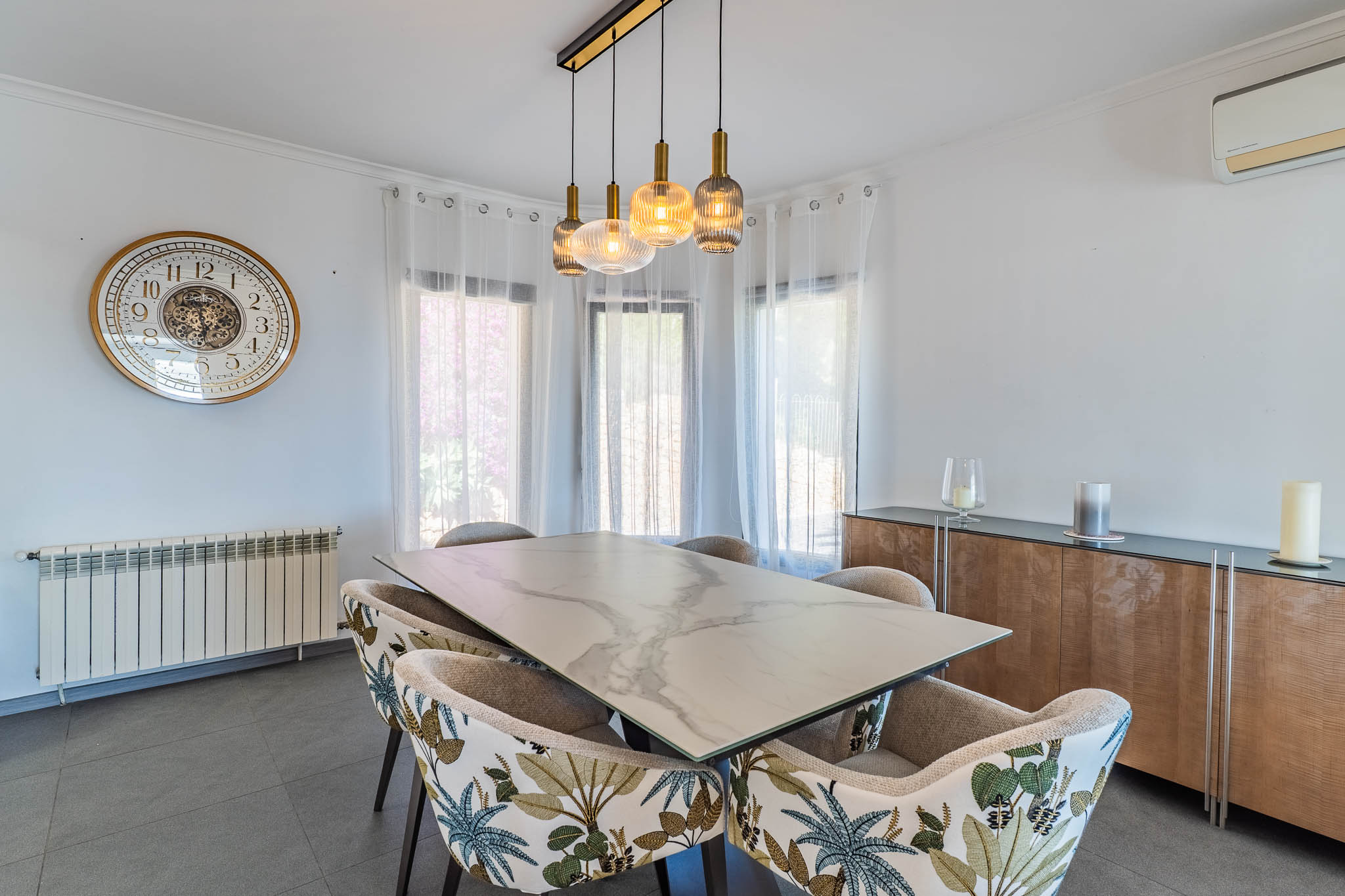 Comedor luminoso con mesa de efecto mármol, sillas de estampado tropical y un moderno candelabro lineal sobre el espacio. Comedor luminoso con mesa de efecto mármol, sillas de estampado tropical y un moderno candelabro lineal sobre el espacio.