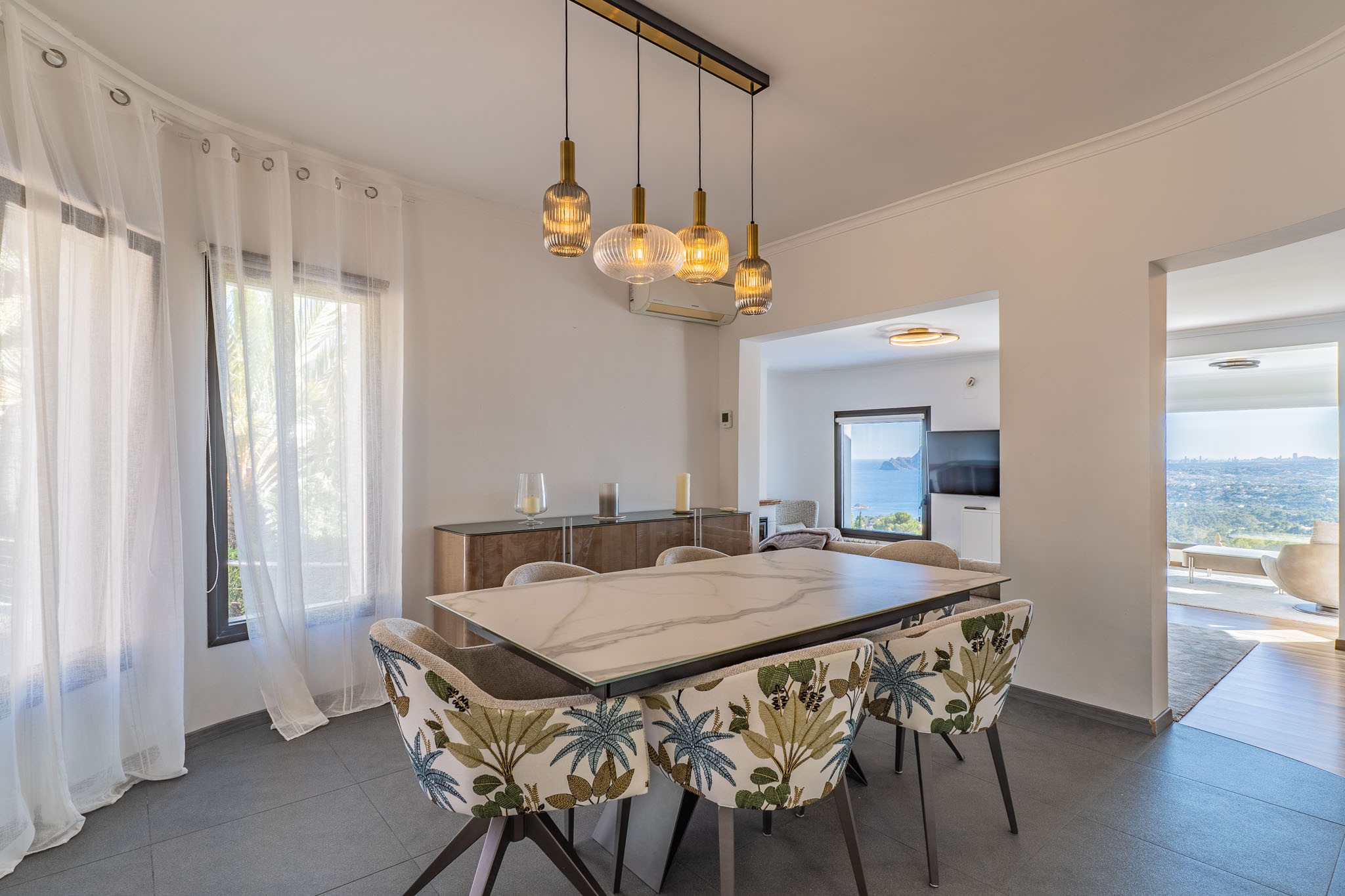 Comedor moderno con mesa de mármol y sillas de diseño tropical. Un candelabro lineal ilumina el espacio, que se abre a vistas panorámicas al mar desde las estancias contiguas. Comedor moderno con mesa de mármol y sillas de diseño tropical. Un candelabro lineal ilumina el espacio, que se abre a vistas panorámicas al mar desde las estancias contiguas.