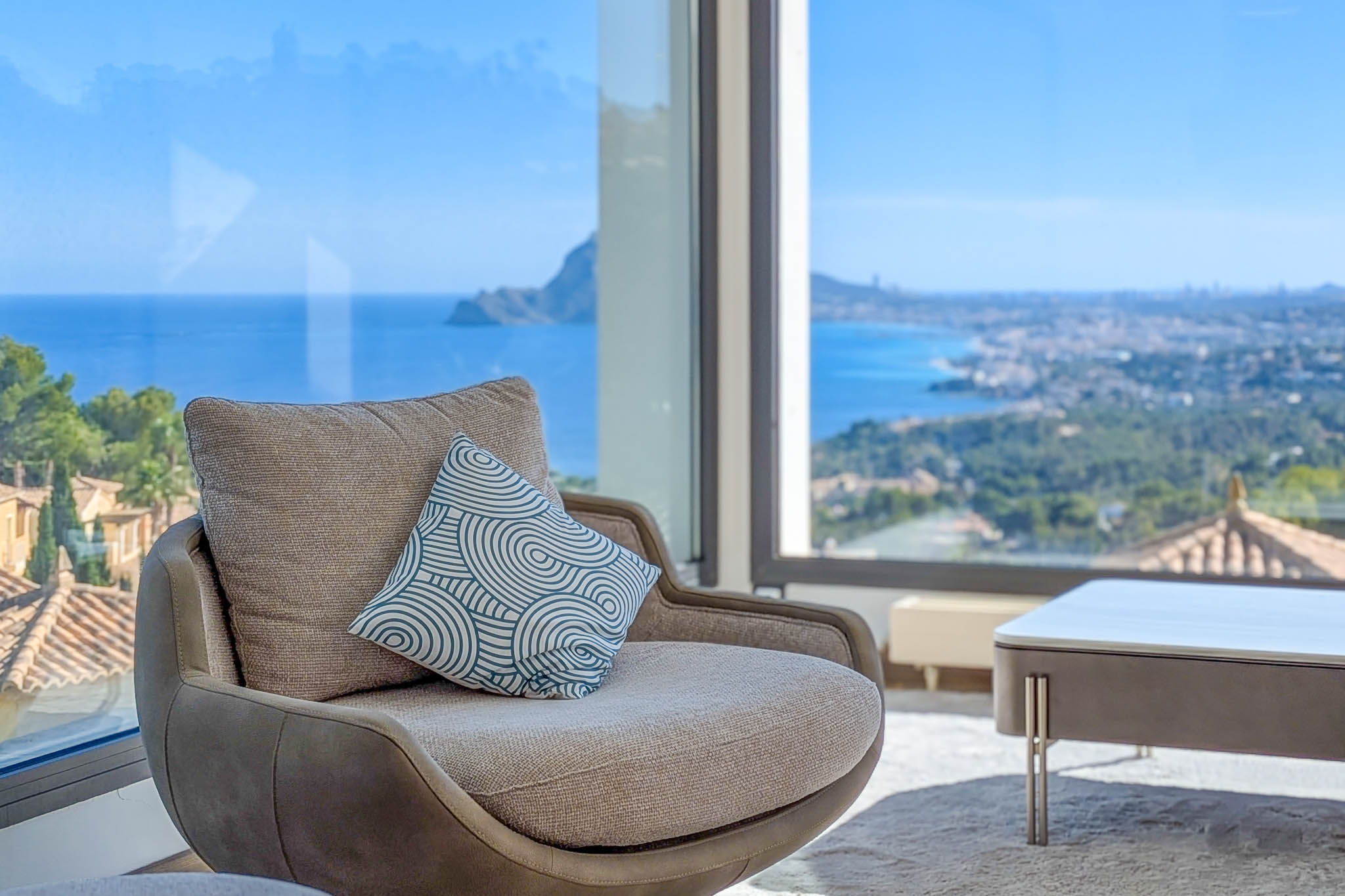 Cómodo sillón junto a ventanal panorámico, ofreciendo vistas ininterrumpidas al mar Mediterráneo y la costa. El cojín decorativo añade un toque moderno al diseño interior. Cómodo sillón junto a ventanal panorámico, ofreciendo vistas ininterrumpidas al mar Mediterráneo y la costa. El cojín decorativo añade un toque moderno al diseño interior.