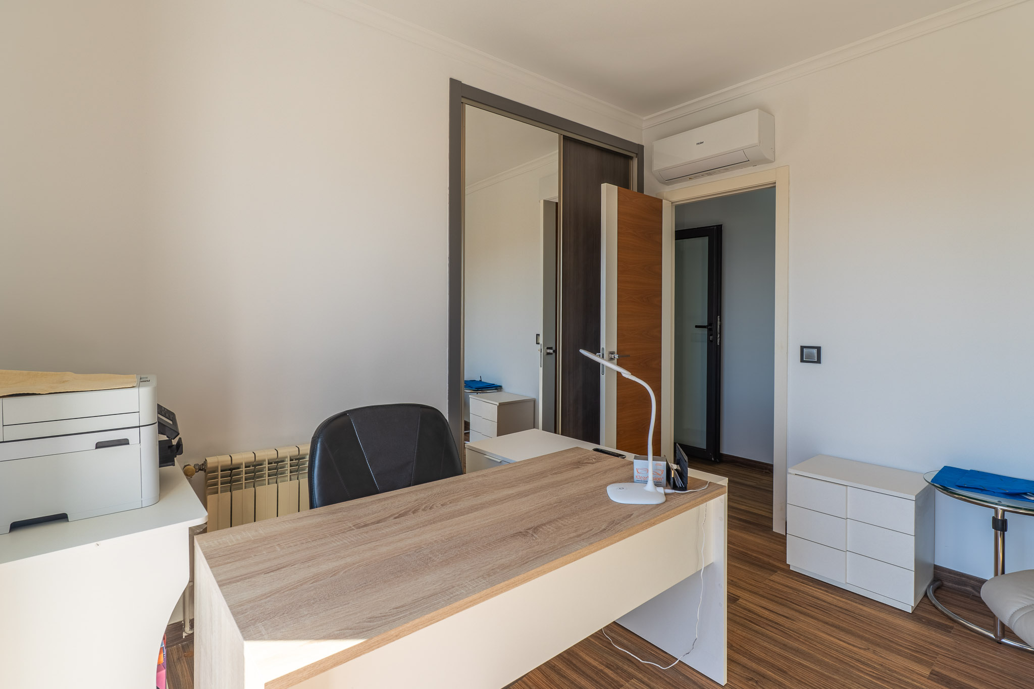Despacho luminoso con escritorio de madera clara, suelo laminado y armario con puertas correderas de espejo, equipado con aire acondicionado. Despacho luminoso con escritorio de madera clara, suelo laminado y armario con puertas correderas de espejo, equipado con aire acondicionado.